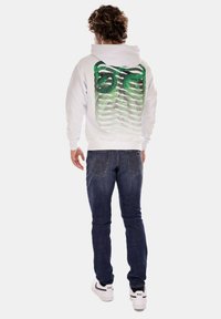 Felpa bianca con un design astratto verde sul retro, abbinata a jeans blu scuro e sneaker bianche. La felpa presenta un cappuccio con stringhe e tasca a marsupio frontale.
