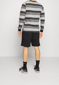 Eine Person trägt schwarze Sportshorts, schwarze Nike-Sneaker, schwarze Socken und ein langärmliges grau-schwarz gestreiftes Shirt und ist von hinten zu sehen.