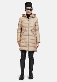 Hellbeige gepolsterter Mantel mit Kapuze, Reißverschluss vorne und zwei seitlichen Taschen. Getragen über schwarzen engen Hosen und knielangen schwarzen Stiefeln.