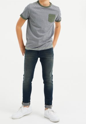 Grijs T-shirt met korte mouwen, groene accenten en een zak, gecombineerd met donkerblauwe slim-fit jeans en witte sneakers. Glad stoftextuur.