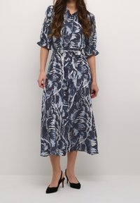 Kvinna iförd en midiklänning i marinblått och vitt med abstrakt mönster, volangärmar, knäppning fram, knytmidja och svarta slingbacks.