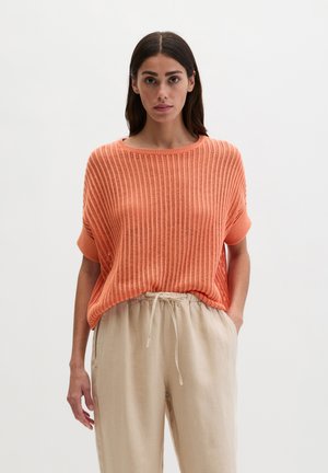 Woman wearing a loose-knit short-sleeve orange sweater tucked into beige drawstring pants, standing with one hand in pocket.