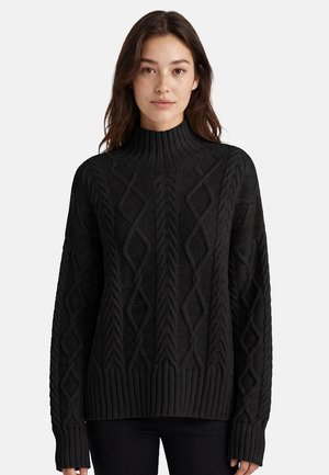 Mujer con un jersey de cuello vuelto negro de punto trenzado con patrones de diamante, posando sobre un fondo claro y liso.