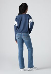 Žena s kitom nosi tamnoplavi duksericu s bijelim prugama, plave traperice i bijele tenisice, stojeći uz jednostavnu sivu pozadinu.