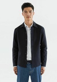 Svart skjorte med knapper laget av teksturert stoff, med spiss krage og lange ermer. Stylet over en hvit rutete skjorte og kombinert med blå jeans.