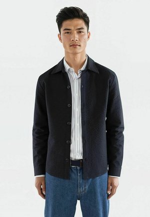 Svart skjorte med knapper laget av teksturert stoff, med spiss krage og lange ermer. Stylet over en hvit rutete skjorte og kombinert med blå jeans.
