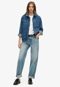 Chaqueta de mezclilla en azul oscuro con cierre de botón, dos bolsillos en el pecho y mangas remangadas, combinada con jeans de corte holgado en azul claro y zapatos negros.