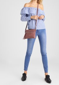 Mujer que lleva una blusa de rayas con hombros al descubierto, jeans ajustados de color azul claro, zapatos negros tipo slip-on y un bolso cruzado marrón.