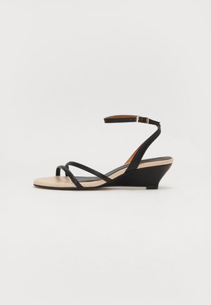 Sandales à talons compensés noires avec un design à lanières, présentant une fine sangle autour de la cheville et une semelle intérieure beige clair. Texture lisse avec des accents minimalistes.