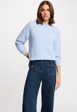 Kvinde iført en lyseblå striktrøje og højtsiddende mørkeblå jeans med frontlommer med klap, stående med den ene hånd bag ryggen.