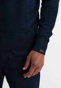 Marineblauwe gebreide trui met een geribde zoom en manchetten, met een label op de manchette. Draagt over donkergekleurde broeken.