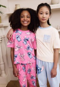 Zwei Kinder posieren zusammen und tragen passende Schlafanzüge. Das eine trägt ein pinkfarbenes Oberteil und eine Hose mit bunten Charaktermotiven; das andere ein cremefarbenes Oberteil und blaue Hosen.