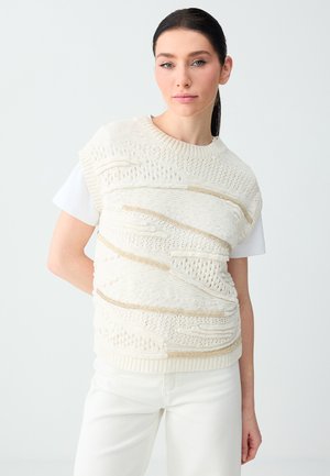 Vrouw met donker haar in een paardenstaart, gekleed in een witte gebreide vest met structuur over een wit shirt met korte mouwen en een witte broek, staand tegen een effen achtergrond.