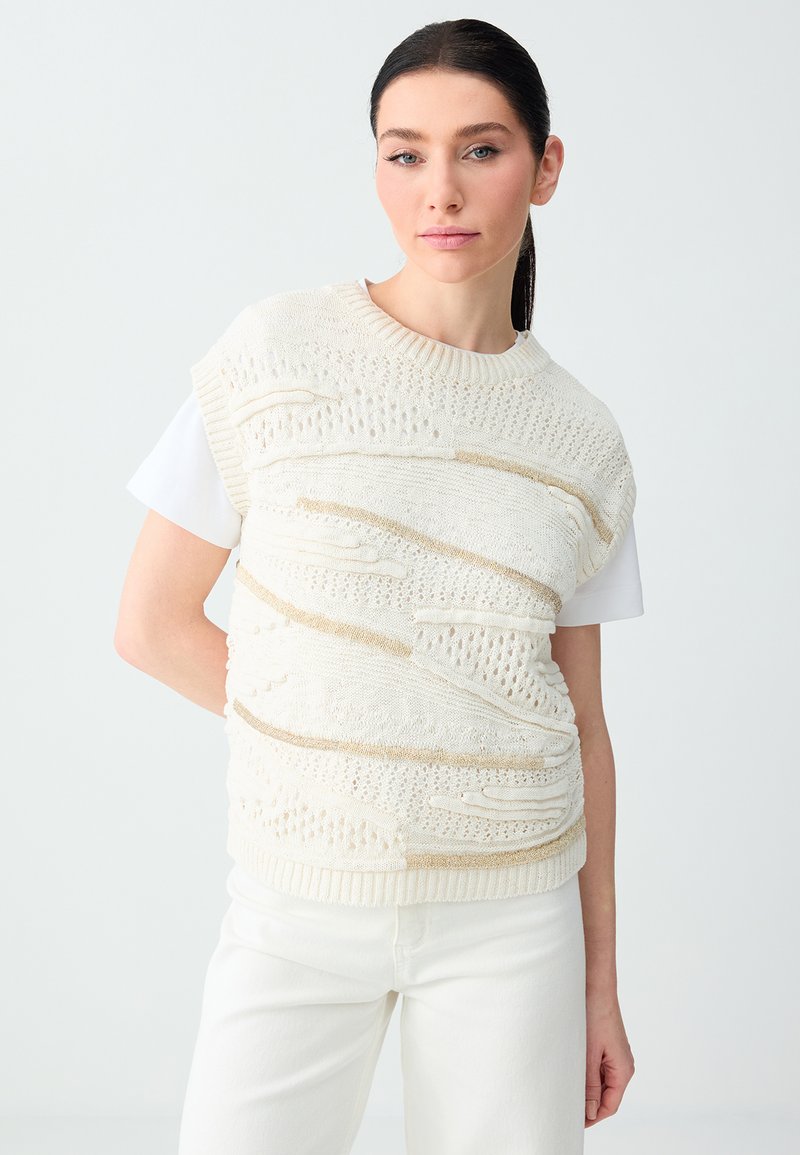 Femme aux cheveux foncés attachés en queue de cheval, portant un gilet en maille texturée blanc par-dessus une chemise blanche à manches courtes et un pantalon blanc, posant devant un fond uni.