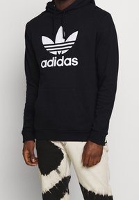 Svart hoodie i bomull med en kenguruficka, med en vit Adidas-logotyp och tre ränder på en bladdesign. Ribbmuddar och nederkant.