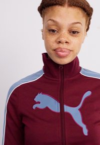 Bordeaux kleurig sportjack met lichtblauwe accenten en een groot Puma-logo op de linkerkant van de borst. Beschikt over een hoge kraag en een ritssluiting aan de voorkant.