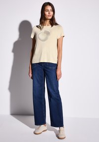 T-shirt beige a maniche corte con un design grafico circolare, abbinato a jeans blu scuro larghi e sneakers di colore chiaro.