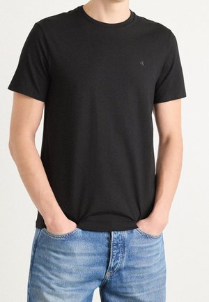 Mężczyzna ubrany w gładki czarny t-shirt z krótkim rękawem z małym logo na piersi oraz niebieskie dżinsy, ręce w kieszeniach.