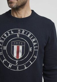 Námořnická modrá mikina vyrobená z bavlny, s kulatou grafikou se štítovým logem, vodorovnými pruhy a rokem "1924" uprostřed.