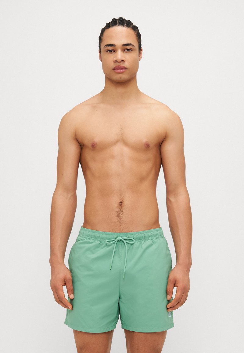 Shorts de baie verzi cu talie elastică și șnur de ajustare. Material neted, lungime până deasupra genunchiului, fără modele sau accente vizibile.