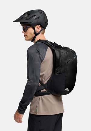Zaino da ciclismo nero con più scomparti, spallacci imbottiti e dettagli riflettenti, indossato sopra una maglietta a maniche lunghe grigia e beige.