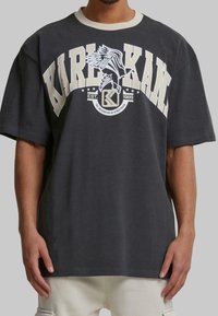 Gray cotton oversized t-shirt with white collar. Front features large cream graphic text "KARL KANI," bird design, and circular emblem.