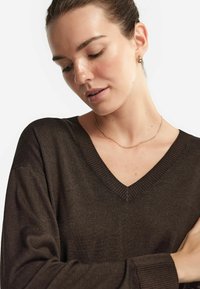 Brauner V-Ausschnitt-Pullover mit gerippten Details am Halsausschnitt und an den Bündchen, mit einer weichen Textur. Das Model trägt eine einfache goldene Halskette.