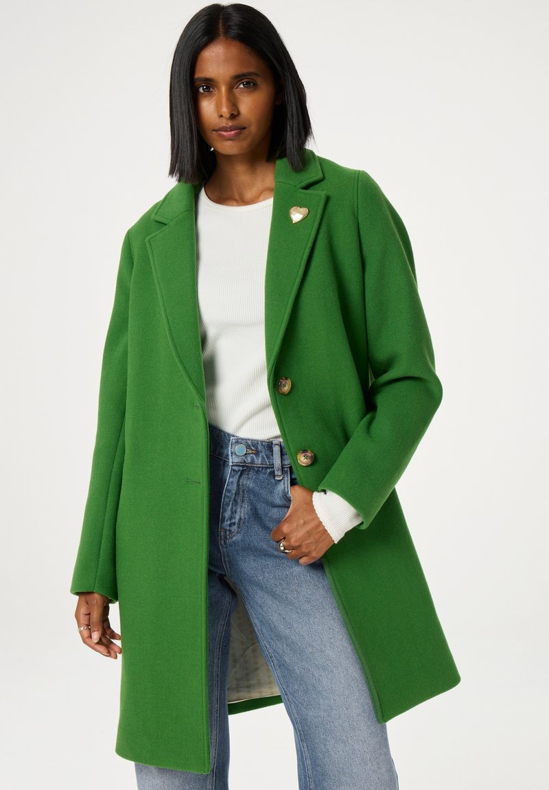 Groene wollen coat met een gekleed revers, twee knopen en een hartvormige broche. Gekoşt over een witte geribde top en blauwe jeans.