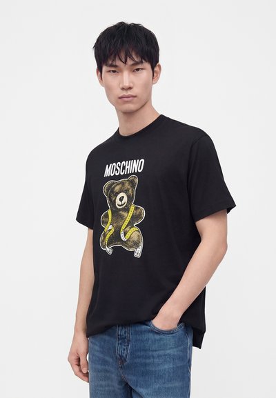 Νέος άνδρας που φοράει μαύρο t-shirt Moschino με μια αρκουδάκι τυλιγμένο με κίτρινη μεζούρα, σε συνδυασμό με μπλε τζιν, στέκεται μπροστά σε λευκό φόντο.