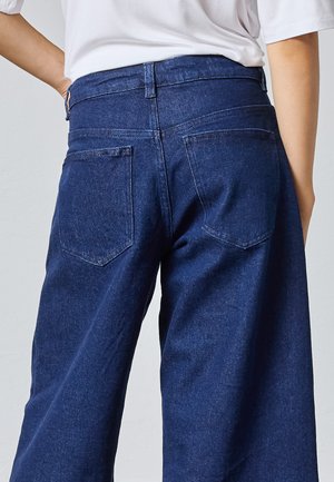 Bagfra af en person iført mørkeblå viddebenede denimjeans og en hvid top med den ene hånd hvilende på hoften.