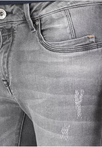 Jeans de mezclilla gris con corte ajustado, que presentan detalles desgastados y un pequeño logo en el bolsillo delantero. Textura y costura sutiles visibles.