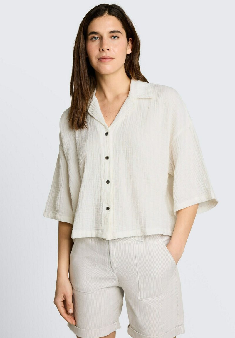 Femme aux cheveux châtains mi-longs portant une blouse blanche ample boutonnée et un short beige clair, une main dans la poche, debout.