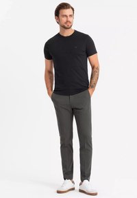 Sort crewneck T-shirt, slim-fit mørkegrå bukser og hvide sneakers med brune såler. Bemærkelsesværdig tatovering på armen, minimal branding synlig.
