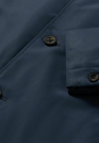 Giacca blu navy con texture liscia, chiusure a bottoni e polsini piegati. Il materiale appare leggero e impermeabile.