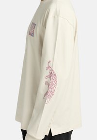 Camicia a maniche lunghe color crema in morbido cotone, con una stampa leopardata rosa sulla manica sinistra e un grafico del sole sul petto.