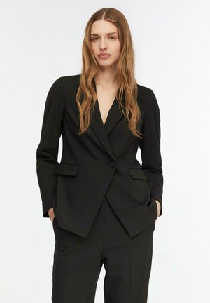 Młoda kobieta z długimi, falującymi włosami, ubrana w dopasowaną czarną marynarkę i pasujące spodnie, ręce w kieszeniach, neutralny wyraz twarzy, jednolite białe tło.