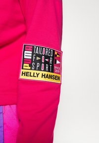 Manga de sudadera de color rosa brillante con una etiqueta rectangular que dice "PUMA TAILORED FOR SPORT SINCE 2019" y "HELLY HANSEN" en texto negro sobre fondo amarillo.