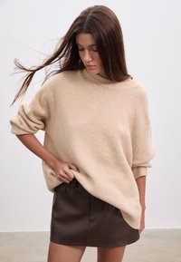 Pull beige surdimensionné en matière douce avec manches retroussées, associé à une jupe moulante en similicuir marron. Design simple, sans motifs ni accents.