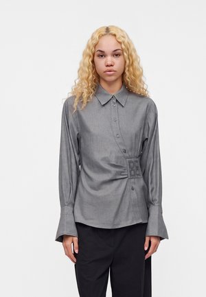 Giovane donna con lunghi capelli ricci biondi, che indossa una camicia grigia con bottoni e dettaglio laterale e pantaloni neri, in piedi su uno sfondo bianco.