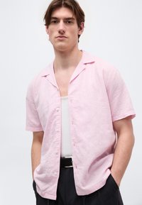 Νεαρός άνδρας που φοράει ανοιχτό ροζ πουκάμισο με κοντά μανίκια πάνω από λευκό αμάνικο φανελάκι και μαύρο παντελόνι, με τα χέρια στις τσέπες, μπροστά από απλό φόντο.