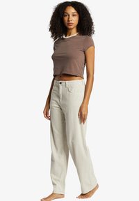 Haut court marron à manches courtes avec un col rond, associé à un pantalon large côtelé beige clair et ample. Pose debout, pieds nus.