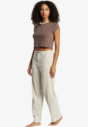 Haut court marron à manches courtes avec un col rond, associé à un pantalon large côtelé beige clair et ample. Pose debout, pieds nus.