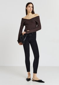Brauner gerippter Crop-Pullover mit schulterfreien Ärmel, schwarze enge Jeans und schwarze Slingback-Ballerinas. Das Model hält eine schwarze Clutch.