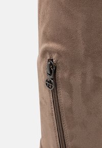 Mochila de ante marrón con una superficie texturizada, herrajes de cremallera negros y un logo circular en acento cerca de la cremallera.