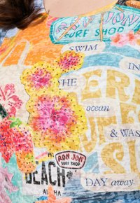 Kleurige korte mouwen blouse met verschillende graphics, bloemdessins in het roze en geel, tekstelementen en strass-accenten op een lichte achtergrond.
