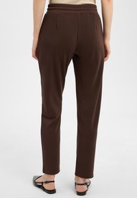 Brązowe spodnie z miękkiego materiału, z elastyczną talią, prostym krojem i mankietami na końcu nogawek. Noszone z czarnymi płaskimi sandałami.