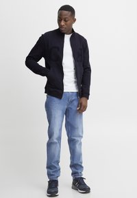 Marinefarbene Zip-Jacke mit "C"- und "H"-Aufnähern, über einem weißen Hemd getragen, kombiniert mit hellblauen Jeans und dunklen Sportschuhen.