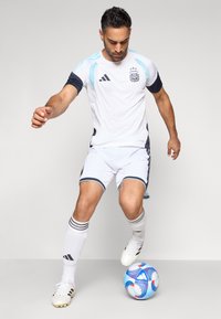 Λευκή και ναυτική ποδοσφαιρική στολή με κοντά μανίκια, που φέρει το λογότυπο AFA, σε συνδυασμό με λευκά παπούτσια και μία πολύχρωμη μπάλα ποδοσφαίρου.