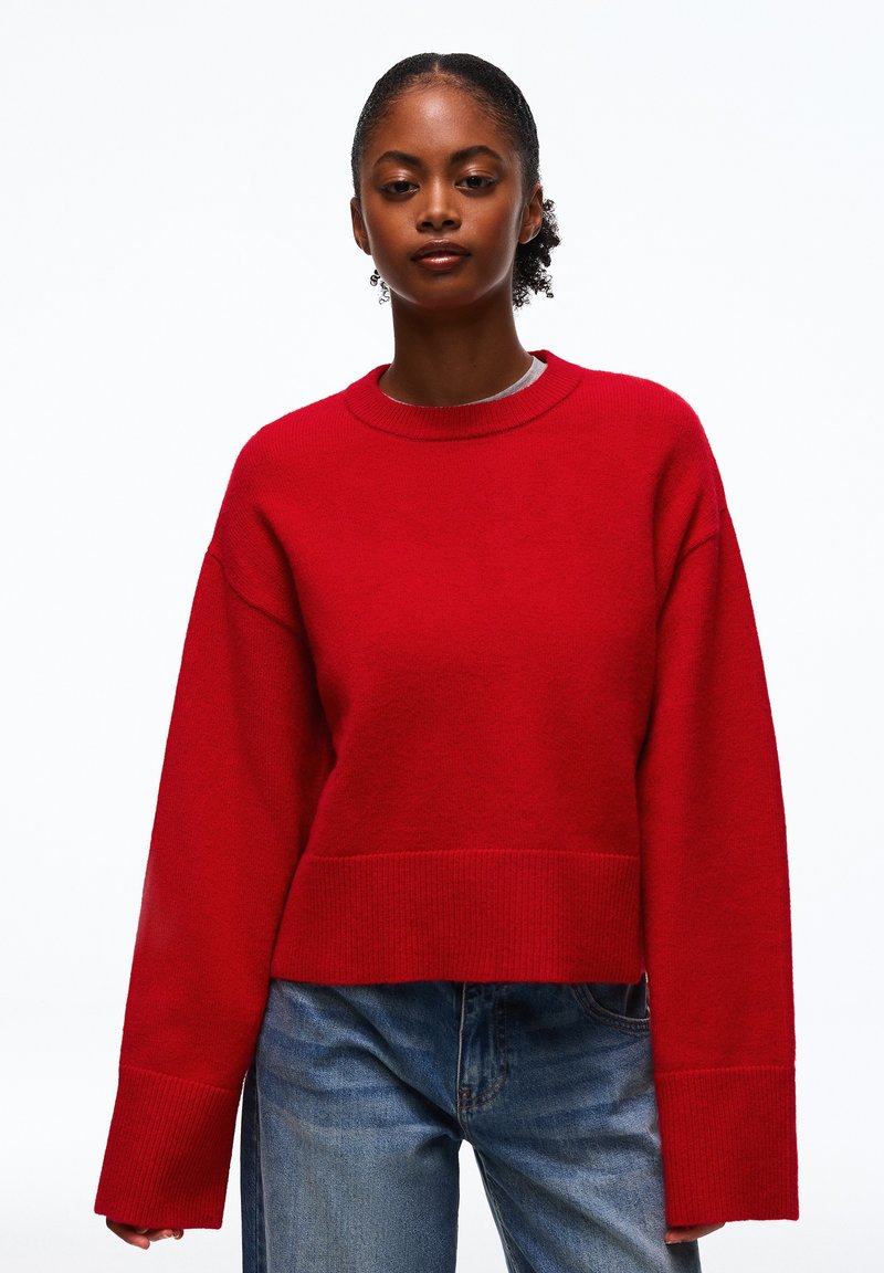 Femme portant un pull rouge à manches longues avec de larges poignets et un jean bleu, debout devant un fond blanc uni.