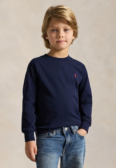 Lyshåret dreng iført en mørkeblå langærmet skjorte med et lille rødt logo og blå jeans, stående med den ene hånd i lommen mod en neutral baggrund.